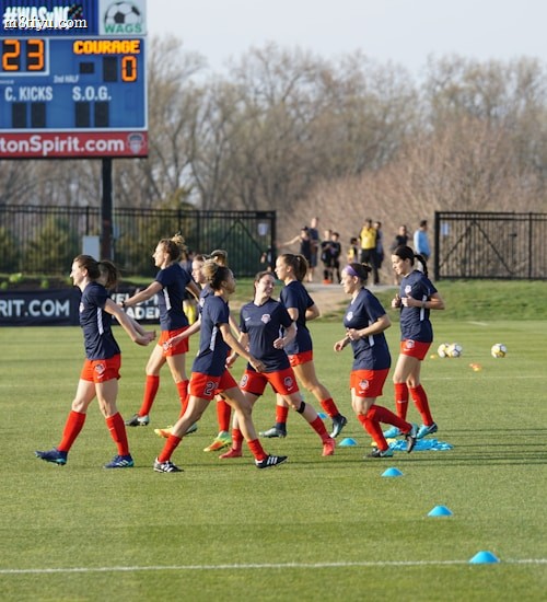 A group of female soccer players in blue jerseys and red shorts are jogging on a grassy field. Training cones are placed on the ground. In the background, a large digital scoreboard shows the time and score. Bare trees and a fence line the field's edge.