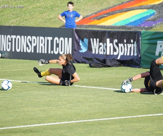 Two football players are performing a training drill on a grassy field. One is leaning back while controlling a soccer ball with her hands, and the other is lying on her back with her legs and arms up, holding another soccer ball. Both are wearing black attire with yellow socks. In the background, banners display social media handles and web addresses, and a person in a blue shirt stands nearby, watching the scene.